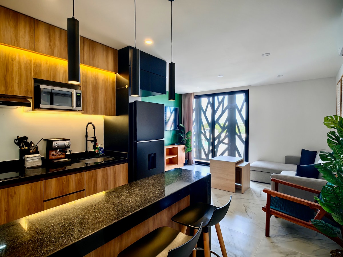 A modern kitchen is featured with sleek cabinetry and dark countertops, complemented by a breakfast bar with three high stools. Natural light fills the living area, which includes a sofa and a minimalist coffee table, set against a backdrop of stylish window designs.