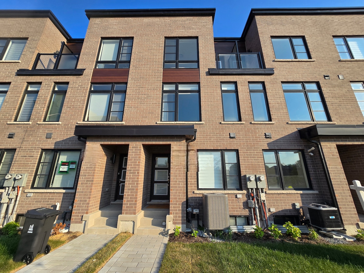 The exterior of a modern townhouse is presented, featuring multiple large windows and a combination of brick and wood accents. A well-paved walkway leads to the entrance, with utility meters visible along the side. Lush greenery borders the base, emphasizing a neat appearance.