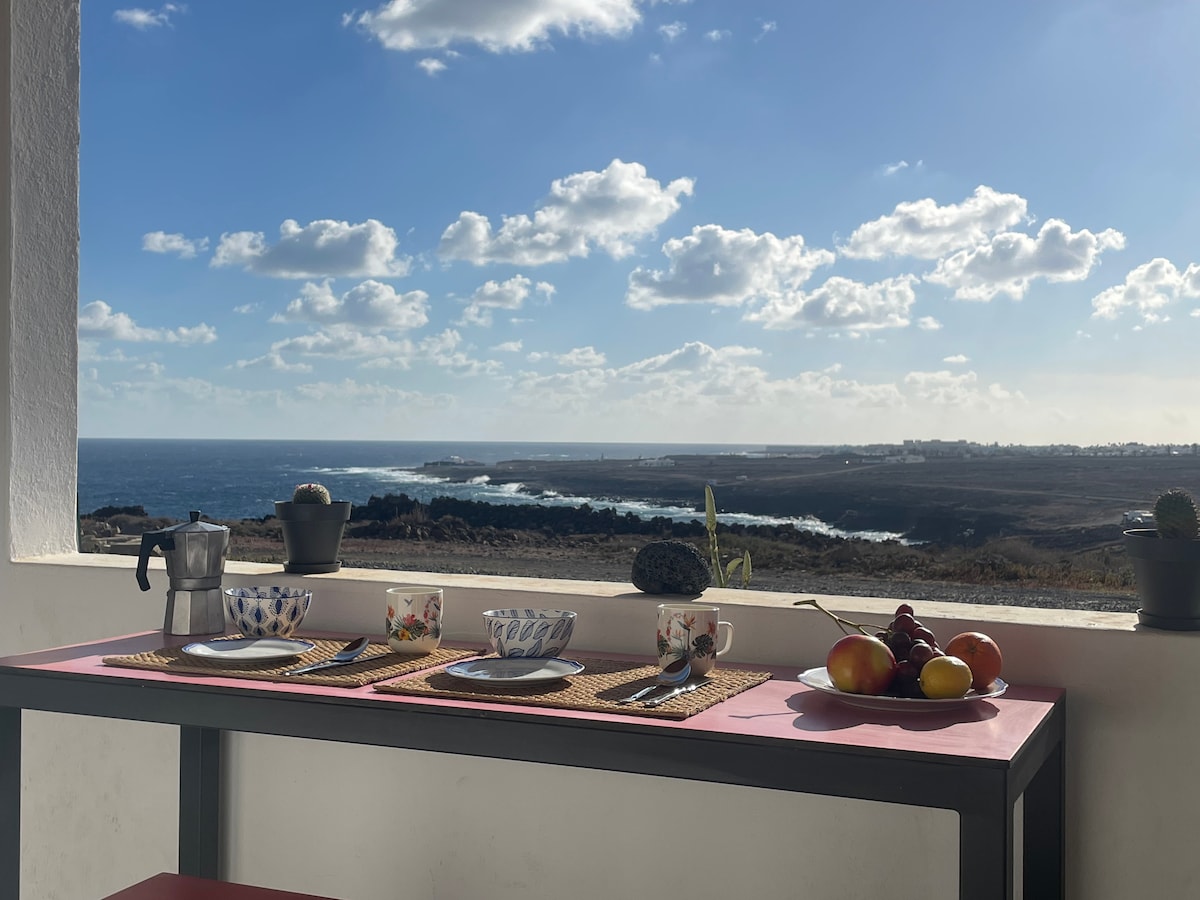 A dining table set for two is positioned on a terrace, offering expansive views of the sea and the coastline. The table features plates, cups, and a coffee maker, with vibrant fruit displayed in a bowl. Soft clouds scatter across a bright sky, enhancing the scene.