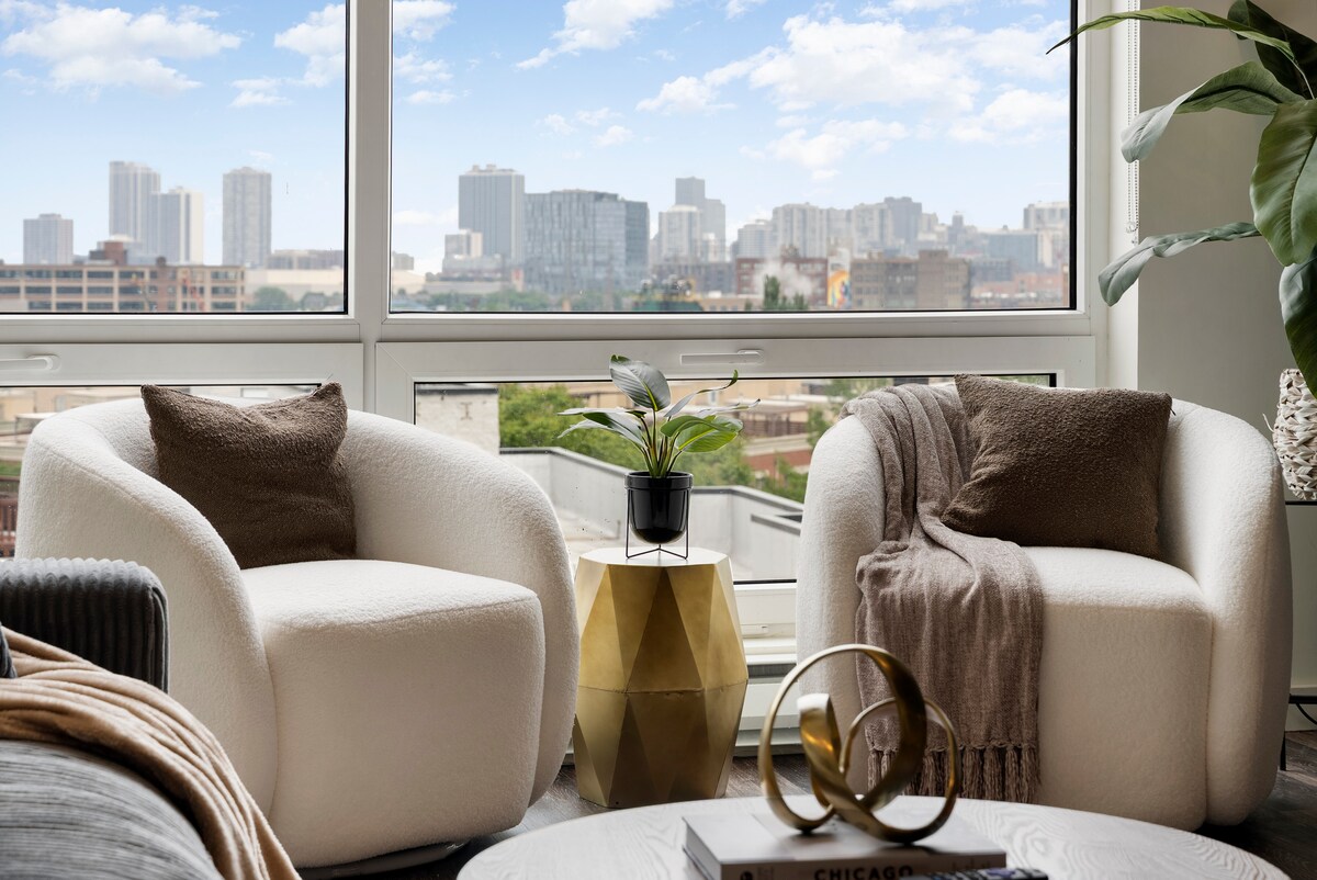 Two plush accent chairs with neutral-toned fabric are positioned near a large window, offering expansive views of the Chicago skyline. A small potted plant sits atop a gold accent table, enhancing the modern aesthetic. Light flows into the space, creating a bright and airy feel.