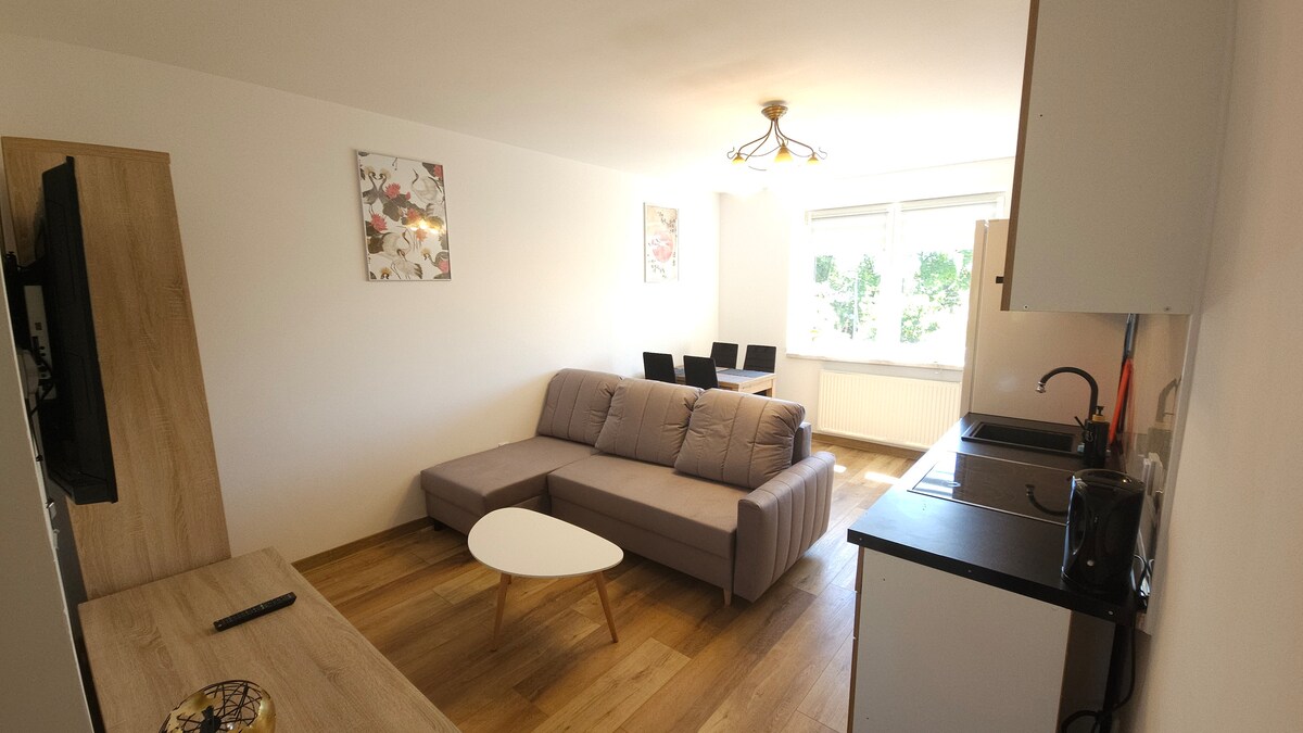 A modern living room features a light-colored sofa set against a neutral wall. A low coffee table is positioned in front, with a television mounted nearby. A dining area with four black chairs is visible, accompanied by a window allowing natural light to brighten the space.