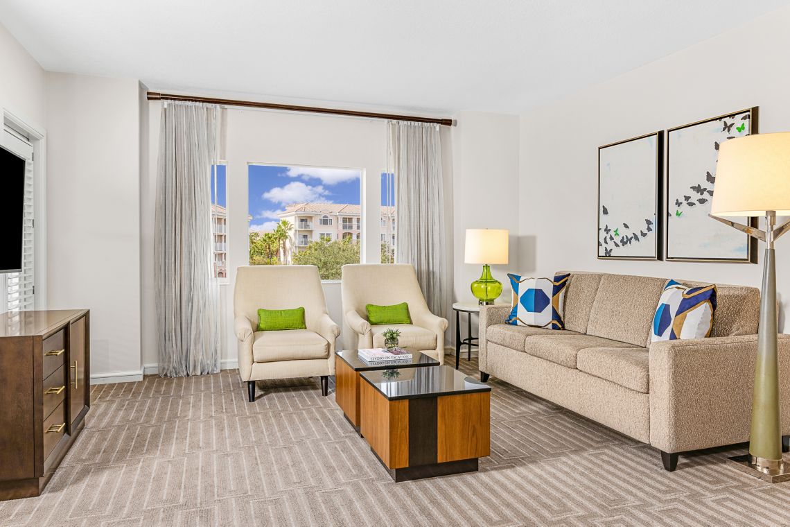 A living area is presented with a light-toned sofa and two matching armchairs. A coffee table with a dark top is centered between the seating. Large windows provide natural light, with partially sheer curtains. Modern artwork complements the decor, creating a relaxed space.