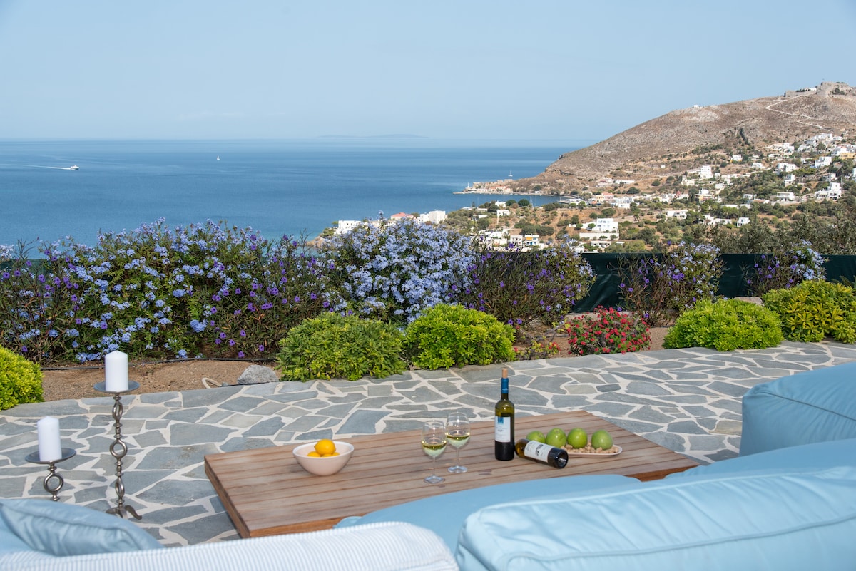 A scenic outdoor seating area features a low wooden table accompanied by soft seating. Refreshments, including a bottle of wine and fruit, are thoughtfully arranged on the table. Vibrant flowers and lush greenery surround the space, with panoramic views of the sea and hills in the background.