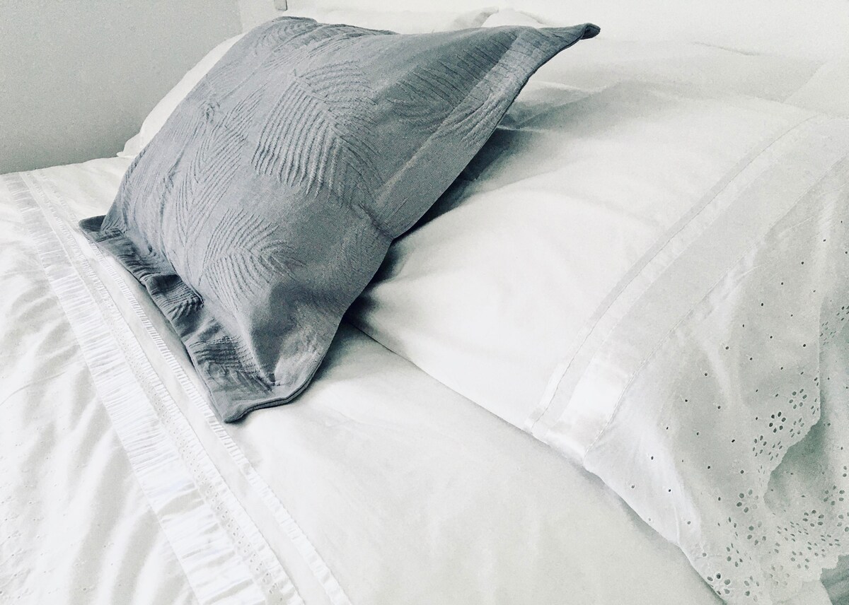 A spacious bed is adorned with crisp white linens and an elegant gray pillow. The bedding features subtle textures and patterns, creating a serene sleeping environment. Soft natural light illuminates the area, enhancing the room's calm ambiance.