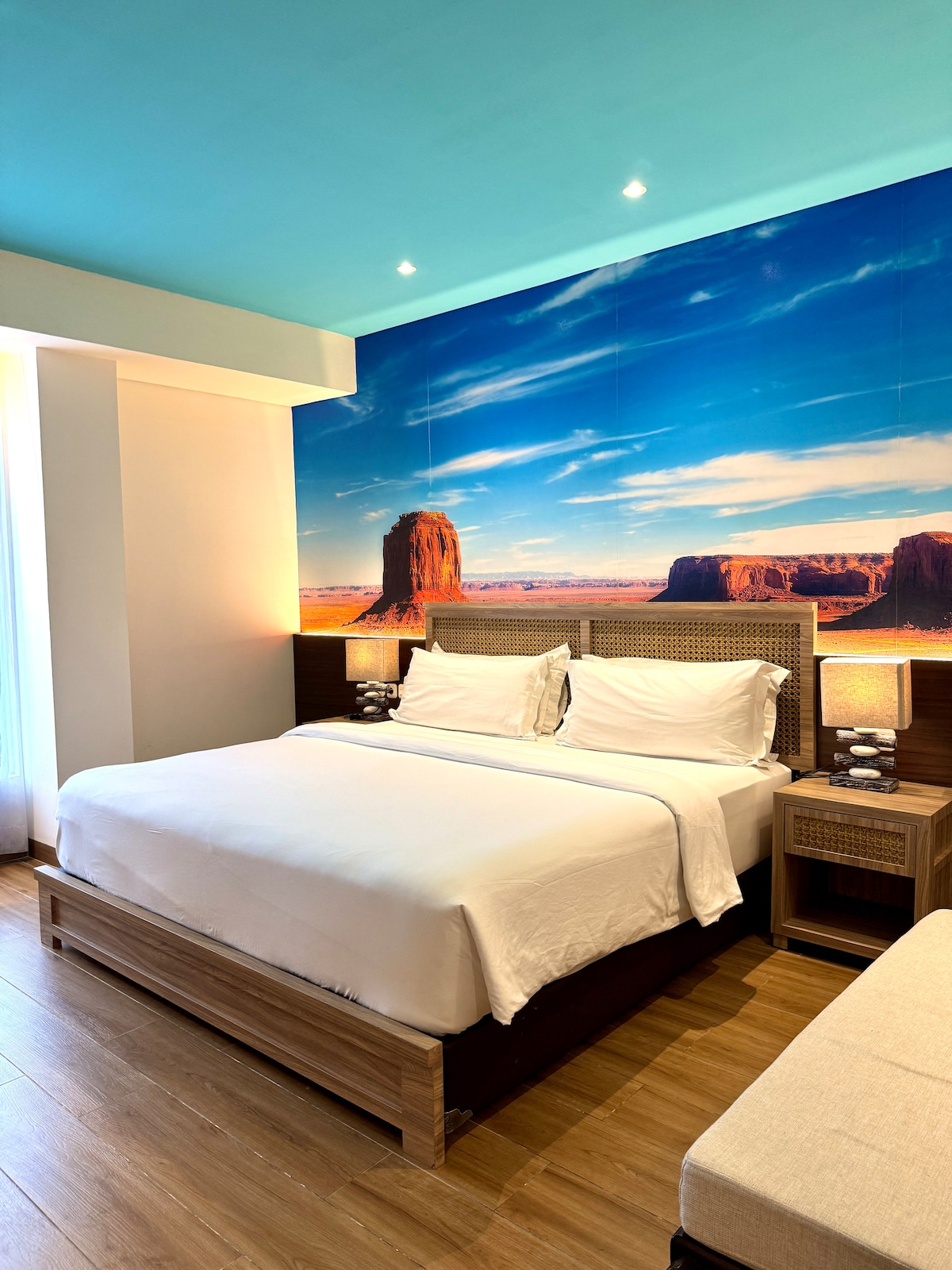 A thematic room is highlighted by a large mural of a desert landscape featuring rock formations. A spacious bed with crisp white linens is centered against the backdrop. Soft lighting is provided by two bedside lamps, and a comfortable seating area is visible near the window.