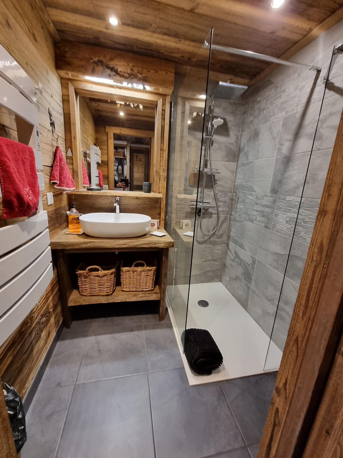 The bathroom features a modern walk-in shower with glass panels, a spacious vessel sink on a wooden countertop, and natural wood accents. Textured tiles line the walls, while dark tiles cover the floor. Baskets are provided for storage, adding a rustic touch.