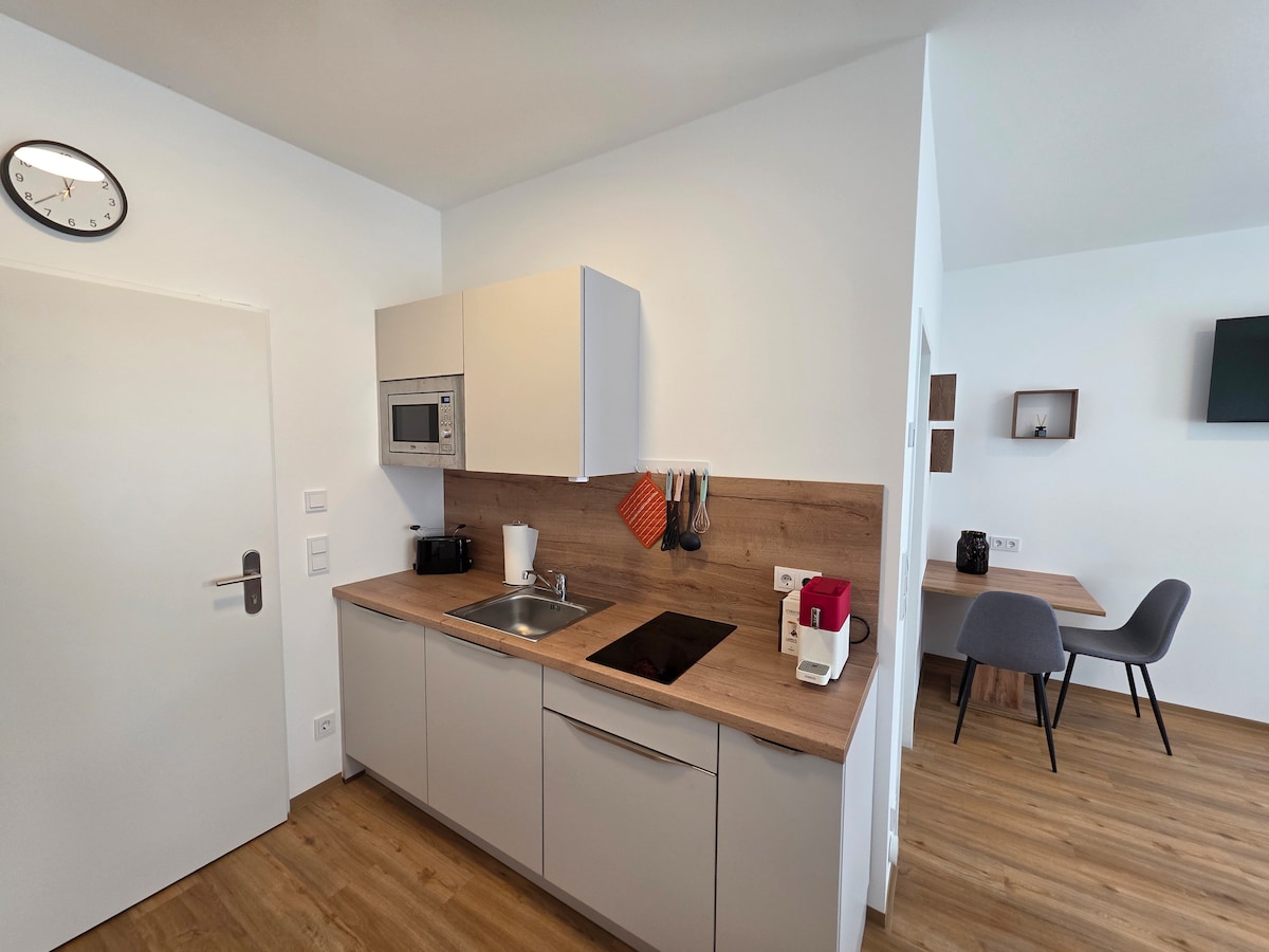 A modern kitchen area is equipped with a countertop, a sink, and a built-in stove. A microwave is mounted above the counter. Kitchen utensils are neatly arranged on the wall. A dining table and chairs are visible in the adjacent area, contributing to a functional space.
