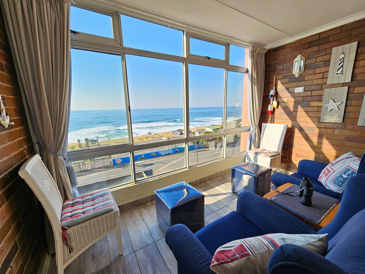A sunlit living area features large windows offering uninterrupted views of the beach and ocean. Two comfortable blue chairs and a wicker chair are arranged invitingly. Light curtains frame the windows, allowing natural light to brighten the space. Decorative nautical elements enhance the coastal ambience.