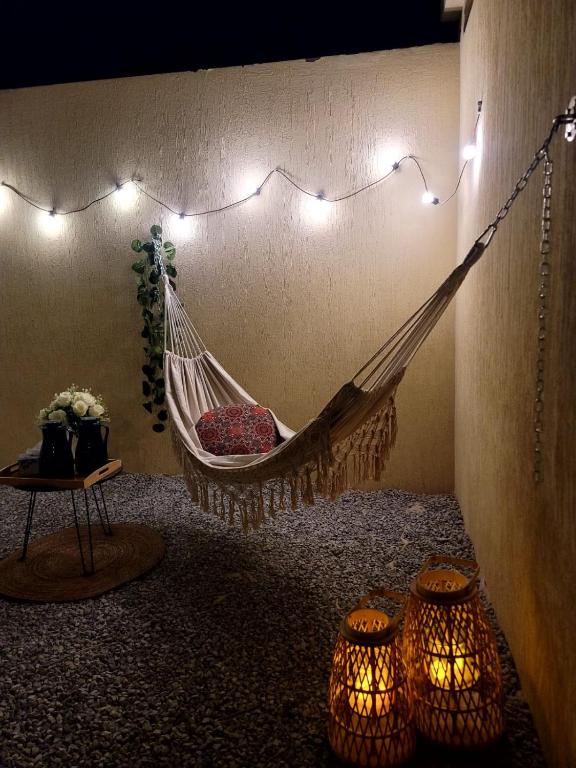 A cozy outdoor area is illuminated by string lights, creating a relaxed ambiance. A hammock with a decorative cushion hangs beside a small round table. Two woven lanterns with soft glow sit on the gravel ground, inviting moments of relaxation.