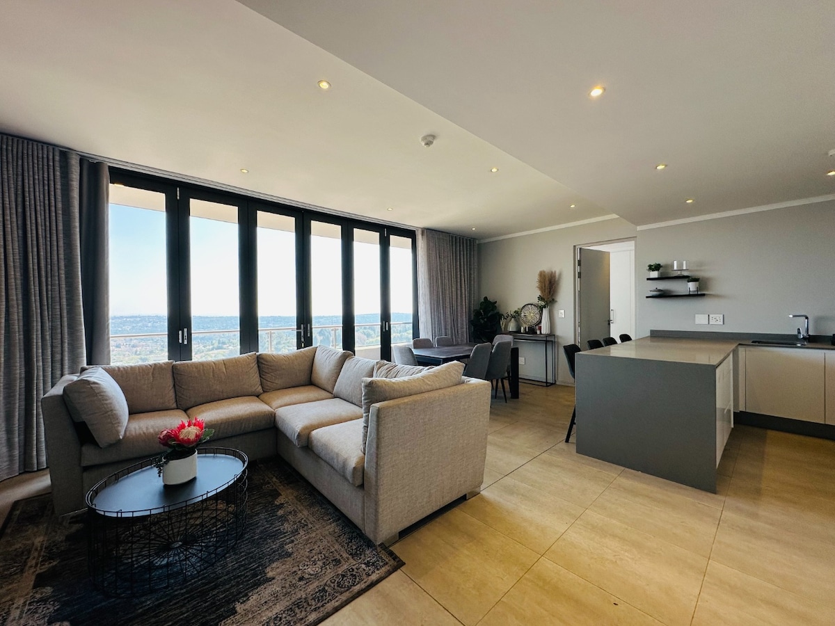 A spacious living area features a large sectional sofa with neutral upholstery and a round coffee table adorned with a small floral arrangement. Expansive windows offer panoramic city views, while a modern kitchen with sleek cabinetry and minimal shelving is visible in the background.