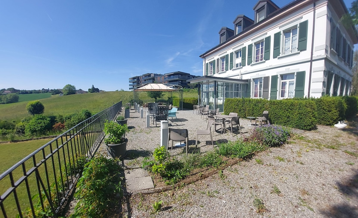 An outdoor terrace is visible, surrounded by a manicured garden and offering a variety of seating options. Sunlight bathes the area, with several tables and chairs placed on gravel. Green hills extend beyond the property, creating a serene backdrop.