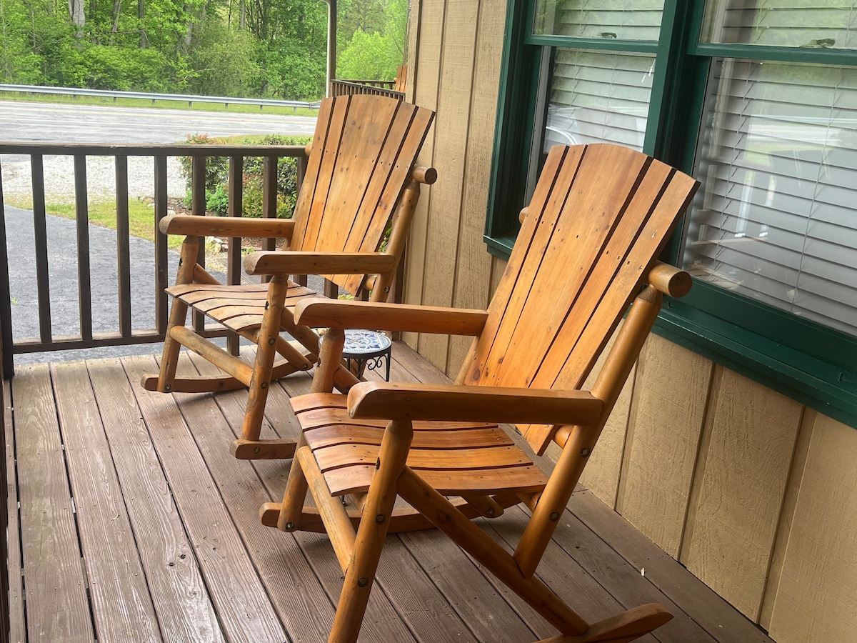 Two wooden rocking chairs are positioned on a covered porch, providing a relaxing spot. The natural wood finish is highlighted by the surrounding greenery. A small table is situated between the chairs, enhancing the inviting outdoor setting.