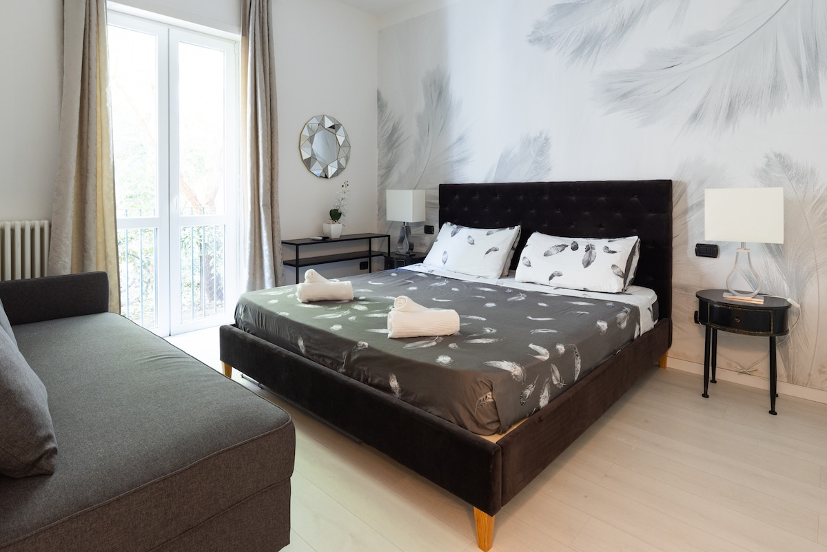A spacious bedroom features a large bed adorned with a patterned duvet and two white towels folded neatly at the foot. A seating area with a gray sofa is visible, alongside a black side table and a mirror reflecting natural light from the window.