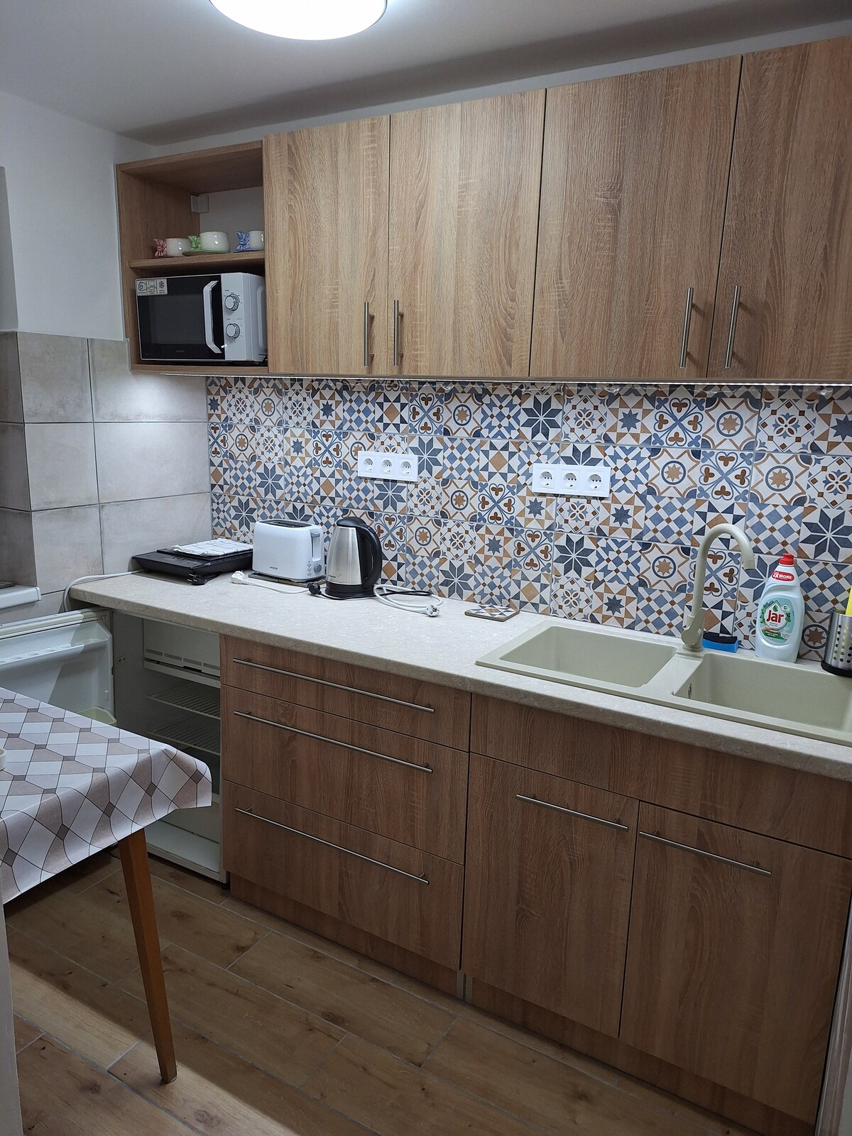 A modern kitchen features wooden cabinetry and a decorative tiled backsplash. Essential appliances, including a microwave, kettle, and a sink, are visible. Counter space allows for meal prep, while a small table adds additional functionality. The kitchen is well-lit with a ceiling fixture.