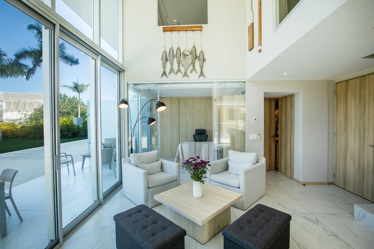 A bright and airy living space features a minimalist design with large glass walls allowing natural light to flood in. Two comfortable armchairs and a sectional sofa surround a wooden coffee table, with decorative items adding a touch of elegance. A small office area is visible in the background.