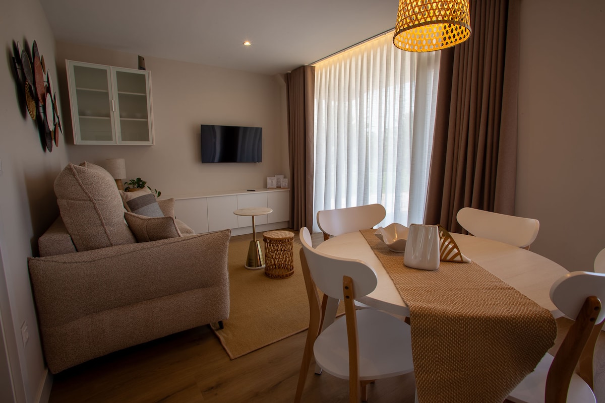 A welcoming living area features a comfortable beige sofa paired with a small circular table. A flat-screen television is mounted on the wall opposite. Natural light filters through sheer curtains, illuminating a dining table set with decorative elements in a neutral-toned setting.