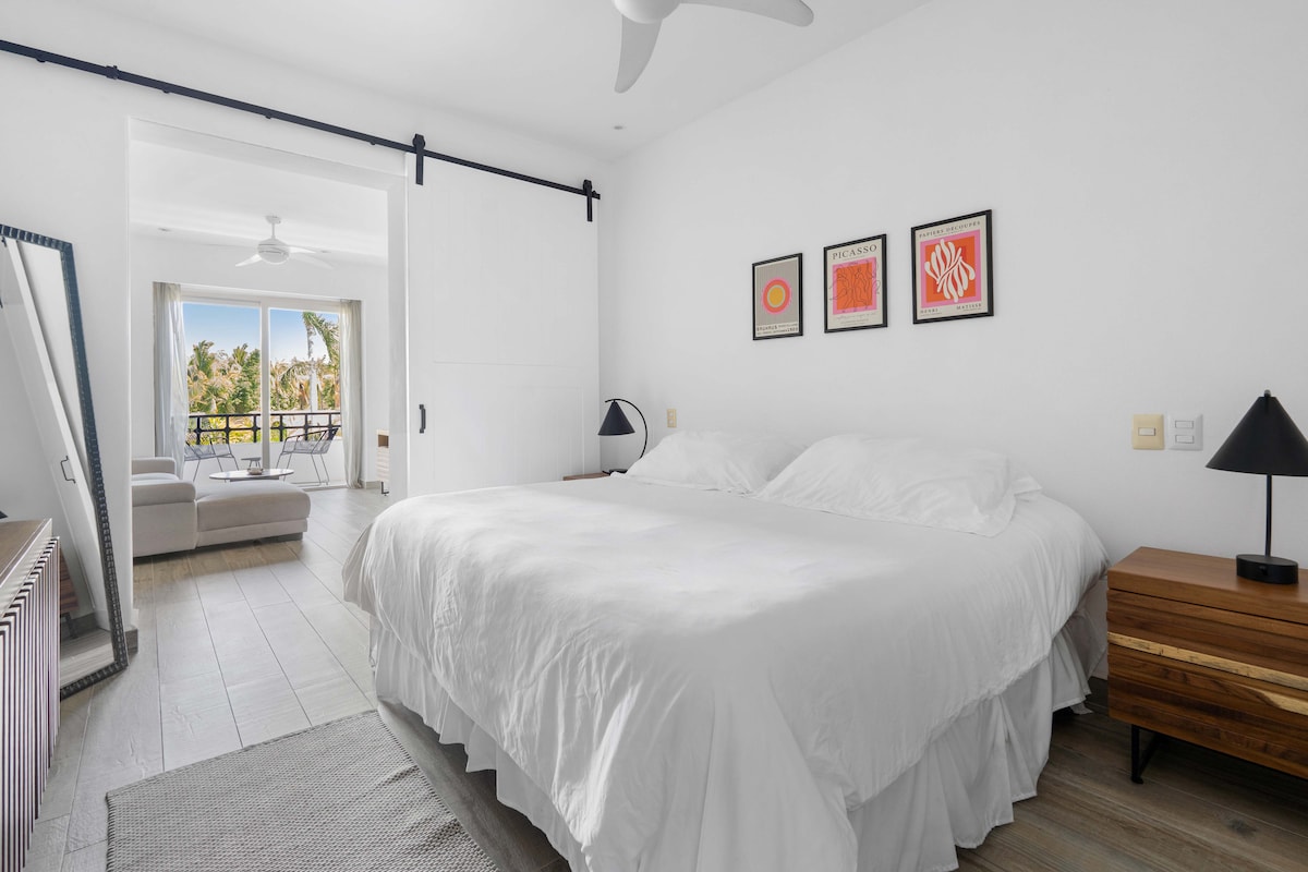 明亮通风的卧室，配有一张加大双人床、白色床单、简约的装饰、带黑色灯具的现代床头柜，以及床上方的艺术画作。 滑动谷仓门通往舒适的起居区，带阳台和热带风光。