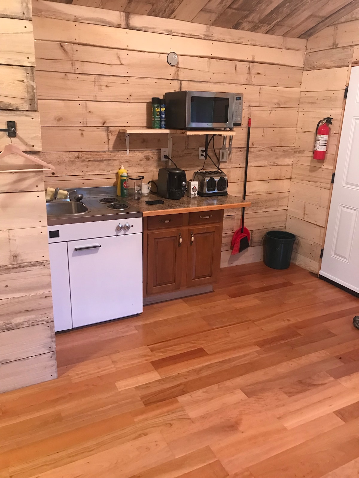 A rustic kitchen area is shown, featuring wooden walls and a warm hardwood floor. Essential appliances include a microwave and a coffee maker. A small wooden cabinet holds condiments, and a countertop provides space for meal preparation. A fire extinguisher is mounted on the wall for safety.