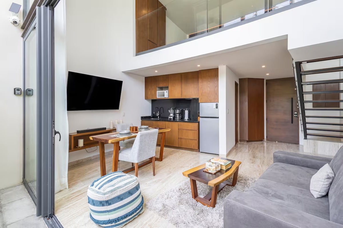 An open living area features a modern grey sofa and a dining table for two. A fully equipped kitchen is displayed with appliances and cabinetry. Large glass doors lead to outdoor views, while the high ceiling enhances the spacious feel of the room.