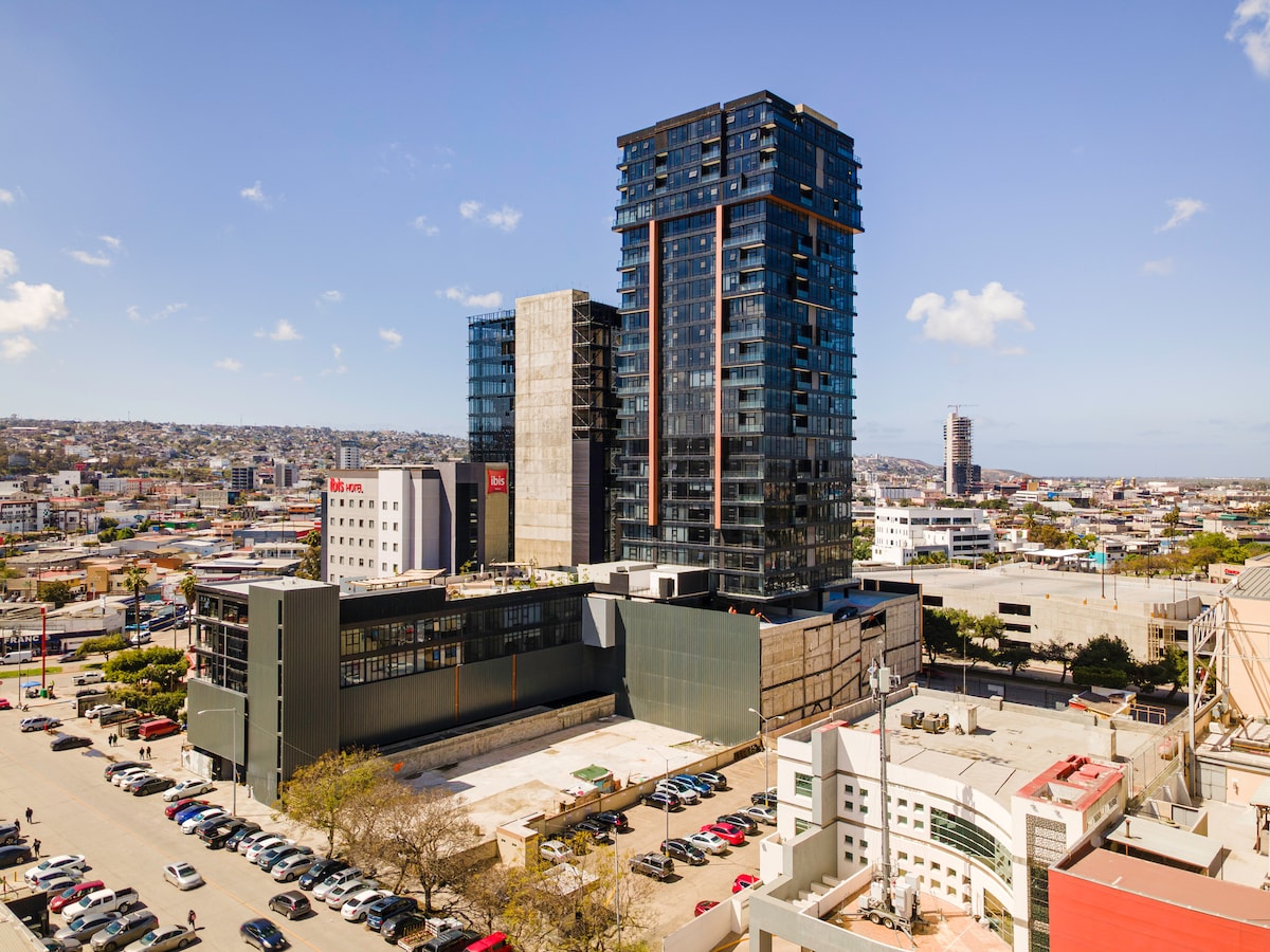 A modern high-rise building is seen in an urban setting, showcasing a mix of glass and concrete architecture. Surrounding areas display a blend of cars parked and city structures, while the horizon features a clear sky, contributing to a sense of spaciousness.