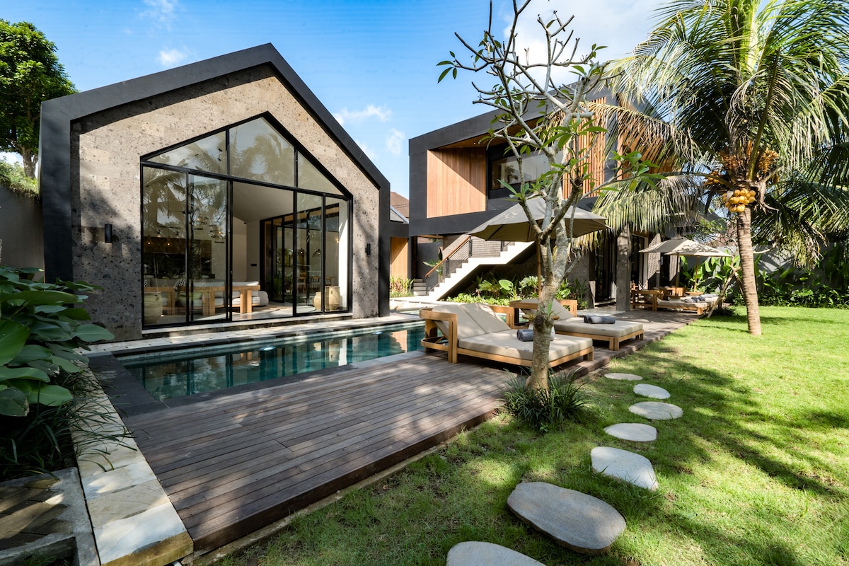 An outdoor view of the villa showcases a blend of modern architecture and lush greenery. Large glass doors create a seamless connection between the indoor and outdoor spaces. A wooden deck surrounds a swimming pool, while sun loungers are positioned on the manicured lawn.