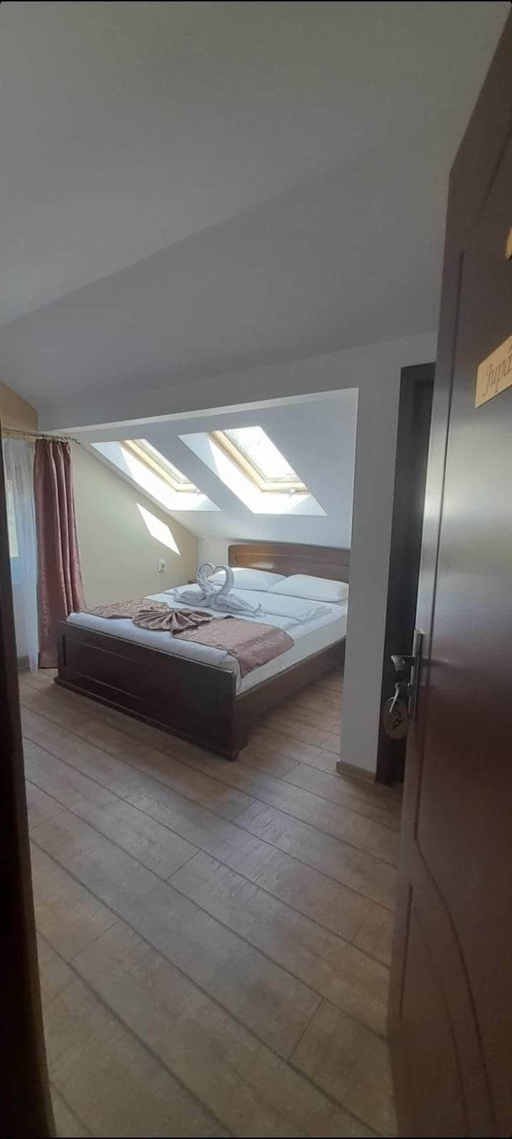 A well-lit room is shown through an open door, featuring a spacious bed with neutral-coloured linens. Natural light filters in through angled skylights above, illuminating the warm wood flooring. Soft curtains frame the window, enhancing the serene ambiance of the space.