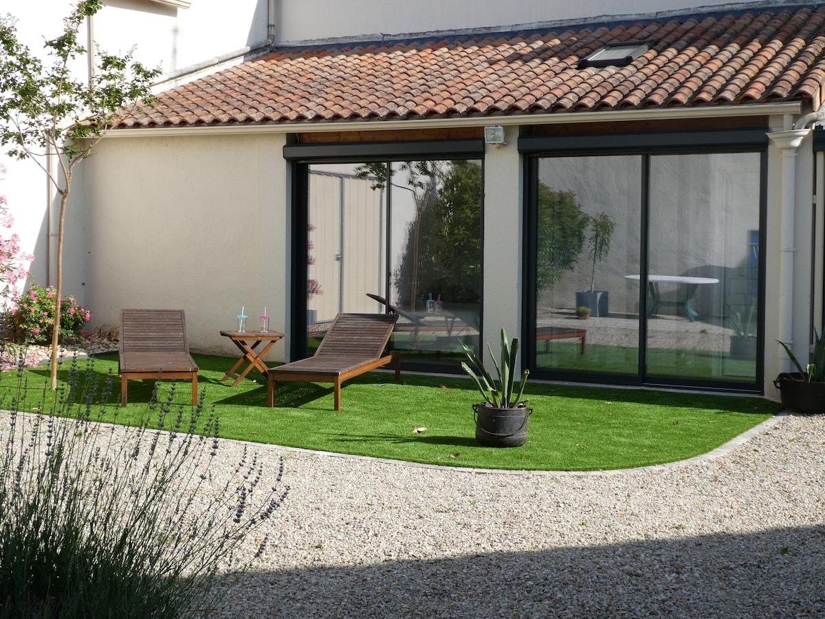 A landscaped outdoor area features two reclining chairs and a small round table, positioned on a grassy surface. Lush green plants are placed in pots nearby, while large glass doors provide a view into the living space. A quiet and tranquil setting is depicted.