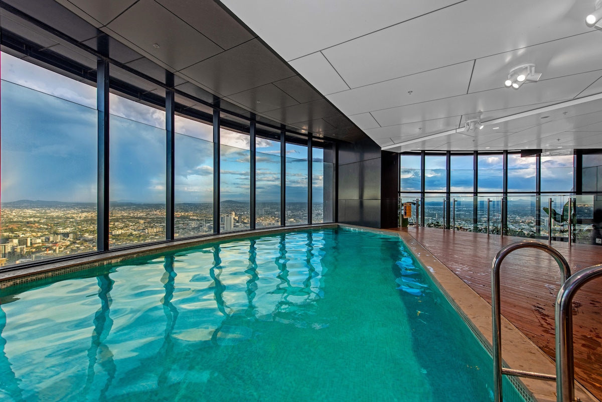 An indoor heated infinity-style pool is visible, offering panoramic views through expansive floor-to-ceiling windows. The serene water reflects the dynamic skyline and distant mountains, while the wooden deck creates a seamless transition to the outdoor space.