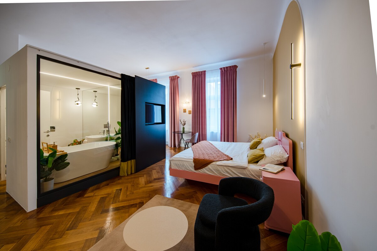 A stylish studio features a spacious sleeping area with a wooden floor and modern décor. A pink bed is set beside large windows dressed with soft curtains. A designated area is seen for relaxation, including a black armchair and decorative plants, with a bathroom visible through a glass partition.