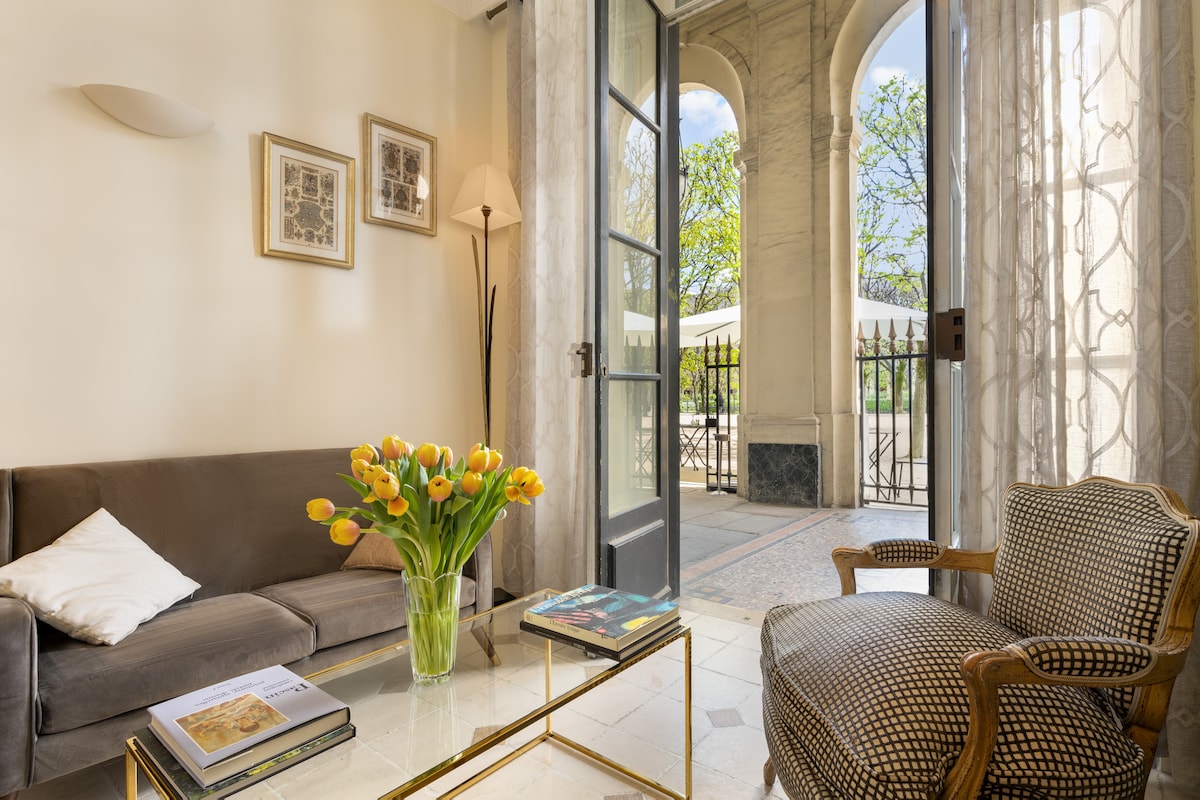 A cozy living room features a gray sofa and a patterned armchair. Bright yellow tulips are displayed in a glass vase on a coffee table. Sunlight streams through floor-to-ceiling French windows, revealing views of an outdoor terrace framed by elegant arches.