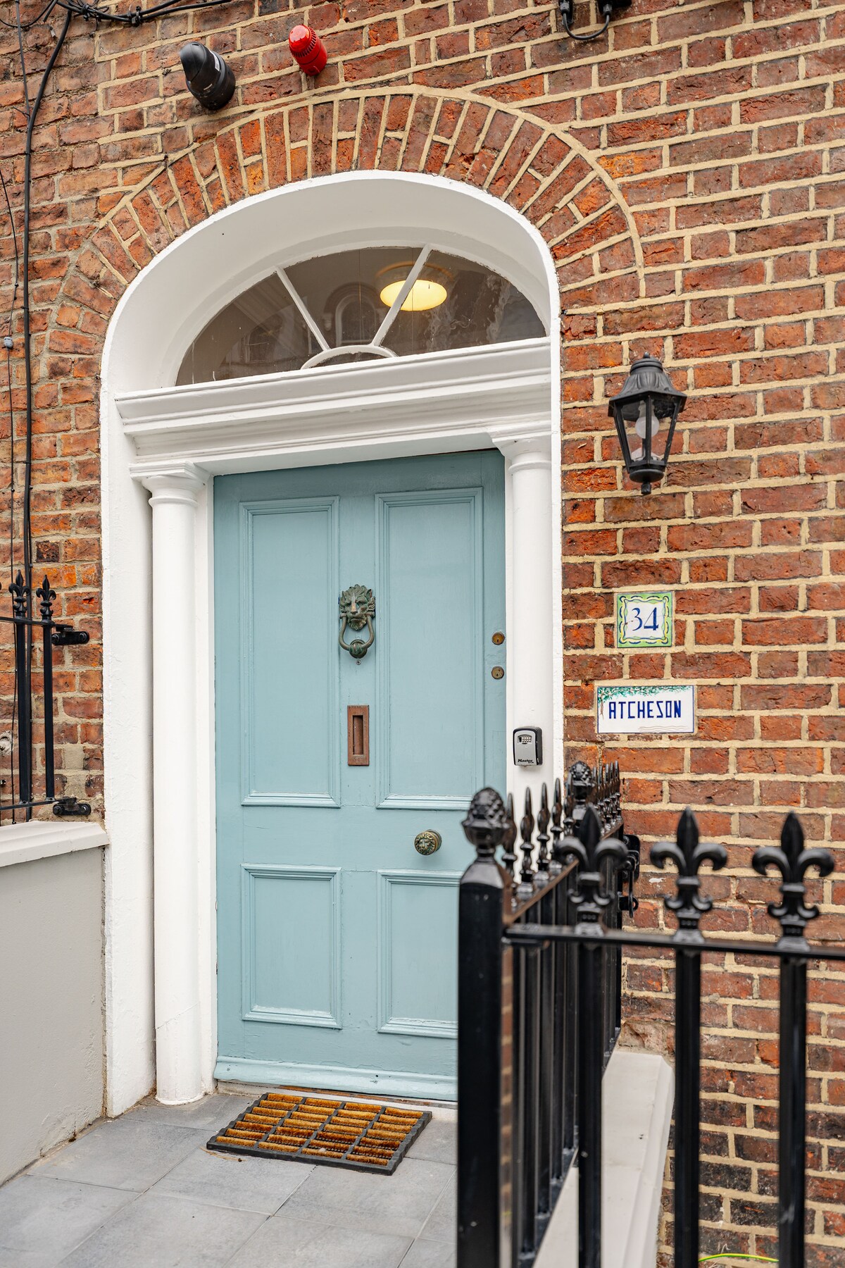 The entrance features a light blue door framed by white columns and a decorative arched window. A brass door knocker and a small doorstep mat are visible. A wrought iron railing runs along the edge of the entryway, enhancing the welcoming appearance of the townhouse.