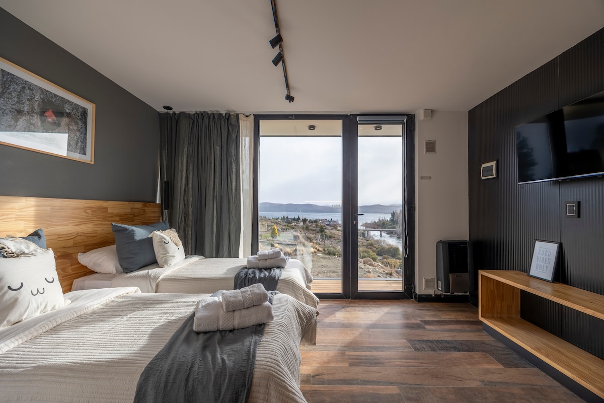 A bright, contemporary room features two spacious beds with soft linens. Large glass doors open to a scenic view of the outdoors. Neutral-toned decor and wooden accents create a calm environment, while a flat-screen TV is mounted on the wall.