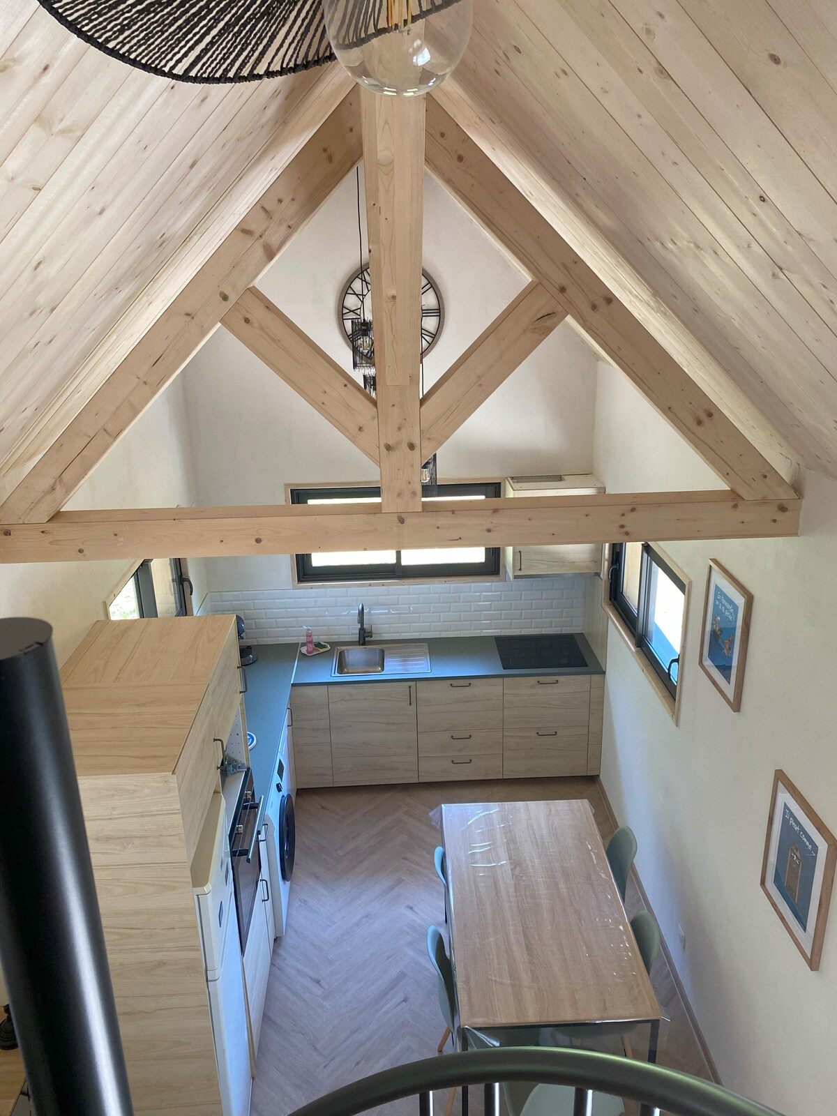 The kitchen area features natural wood elements and modern appliances. A dining table is positioned nearby, surrounded by four chairs. Large windows allow natural light to brighten the space. The high, exposed wooden beams create an airy atmosphere, emphasizing the room's height.