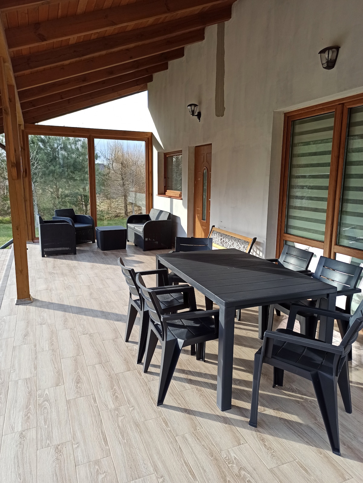 A covered terrace features a dining table surrounded by black chairs. Two lounge chairs are positioned nearby, offering a comfortable seating area. Natural light filters through large windows, and views of the surrounding greenery are visible beyond the terrace.
