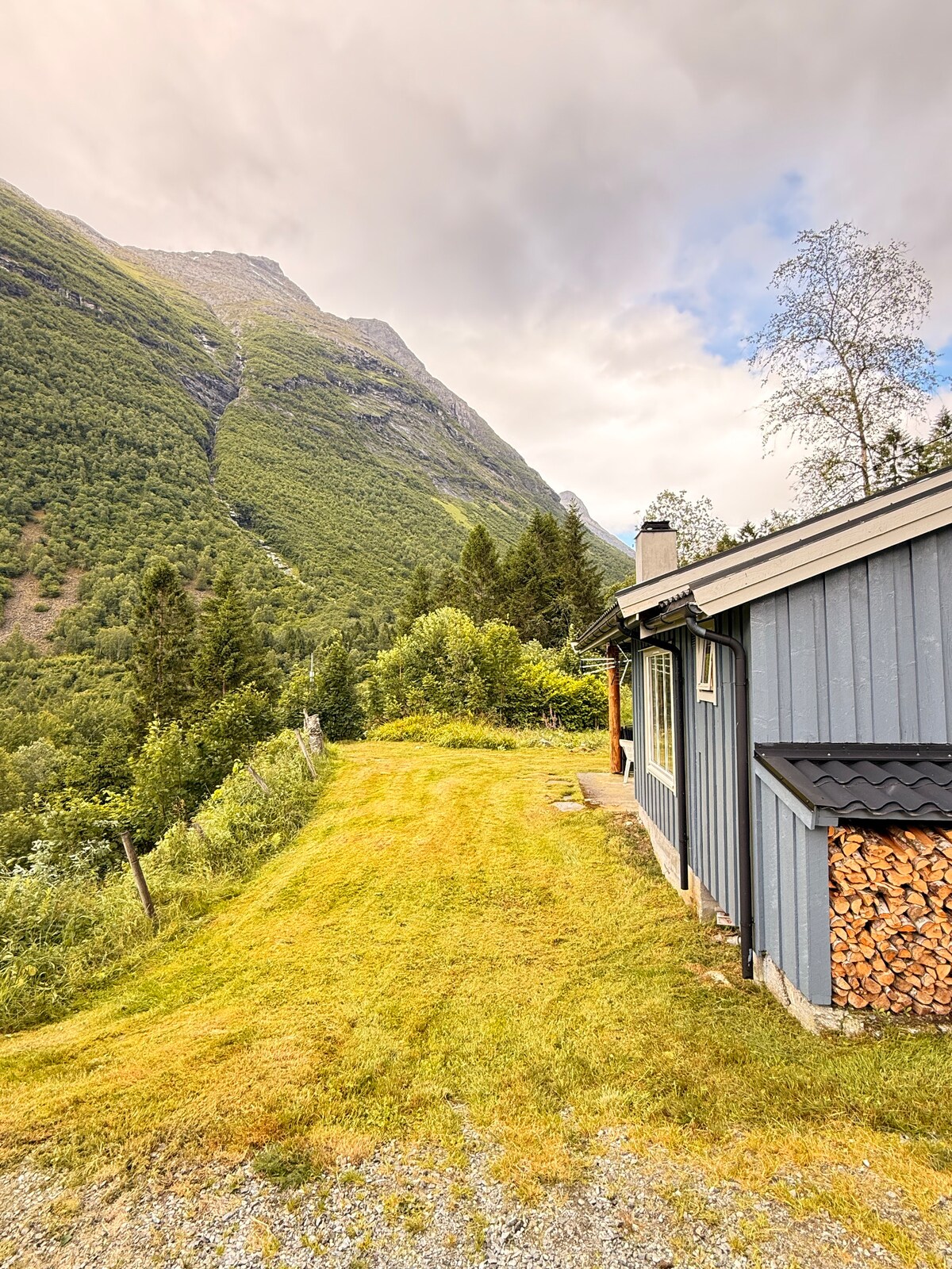 A grassy area leads up to the cozy cabin, framed by lush greenery and steep mountains in the background. Stacks of firewood are neatly arranged beside the cabin, and gentle clouds cover part of the sky, hinting at the natural beauty surrounding the location.