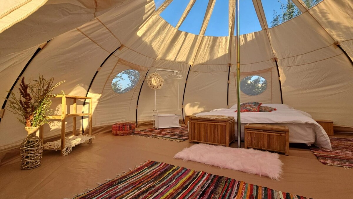 An inviting glamping tent interior features a spacious bed at the center, flanked by wooden side tables. Colorful rugs line the floor, complemented by decor items such as a small clothes rack and woven baskets. Round windows offer natural light and a view of the outdoors.