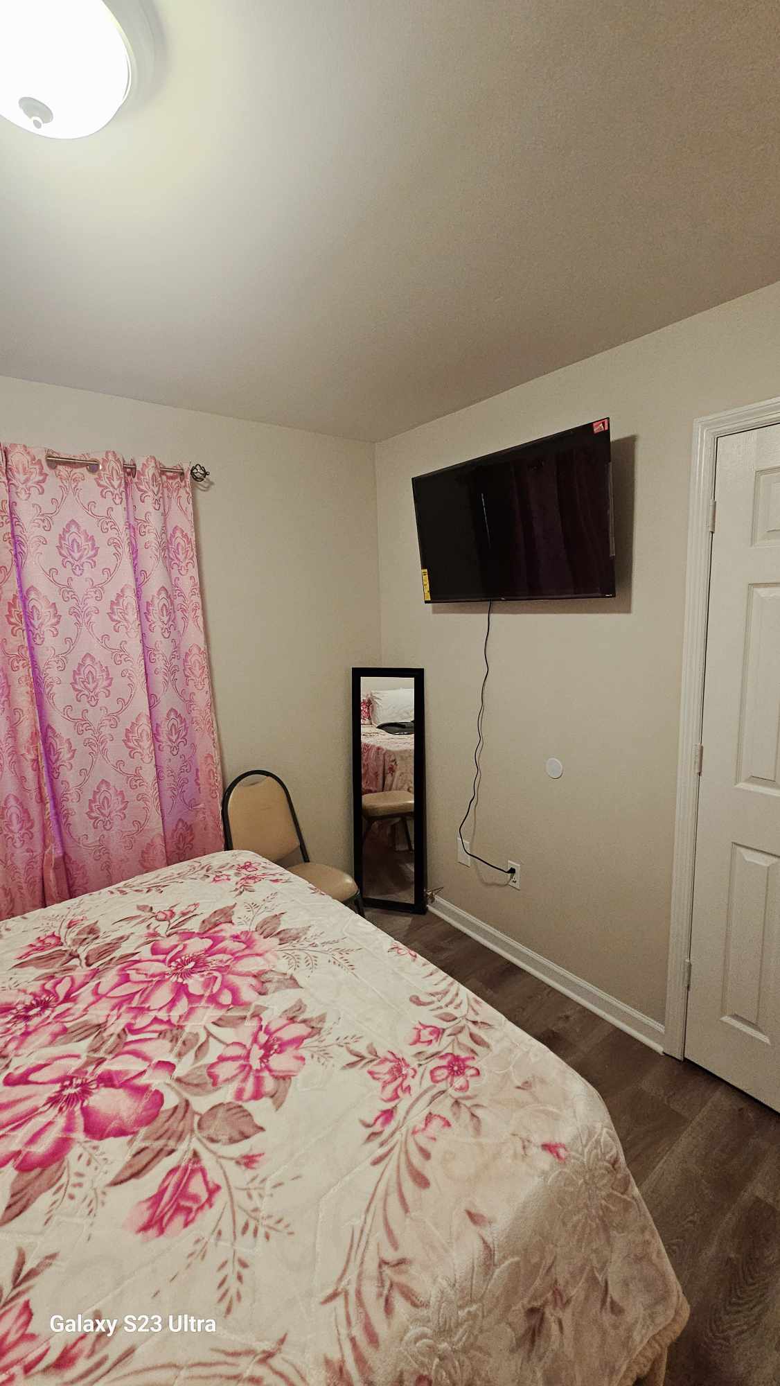 A private room features a comfortable bed with a floral-patterned blanket. A mirror is positioned against the wall beside a chair, while a wall-mounted television is visible across from the bed. Soft pink curtains frame the window, enhancing the room's ambiance.