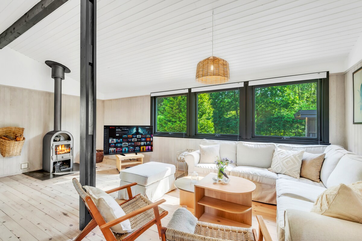A bright living area features large windows offering views of the surrounding greenery. A cozy seating arrangement includes a spacious sectional sofa and wooden chairs. A coffee table sits centrally, while a modern TV and a warm wood-burning stove are positioned against the wall.