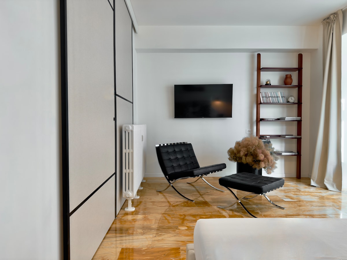 A minimalist living area showcases a sleek design with black lounge chairs positioned elegantly. A mounted flat screen TV is visible, alongside a bookshelf displaying select decor and literature. Natural light filters through a large window, enhancing the warm tones of the polished marble floor.