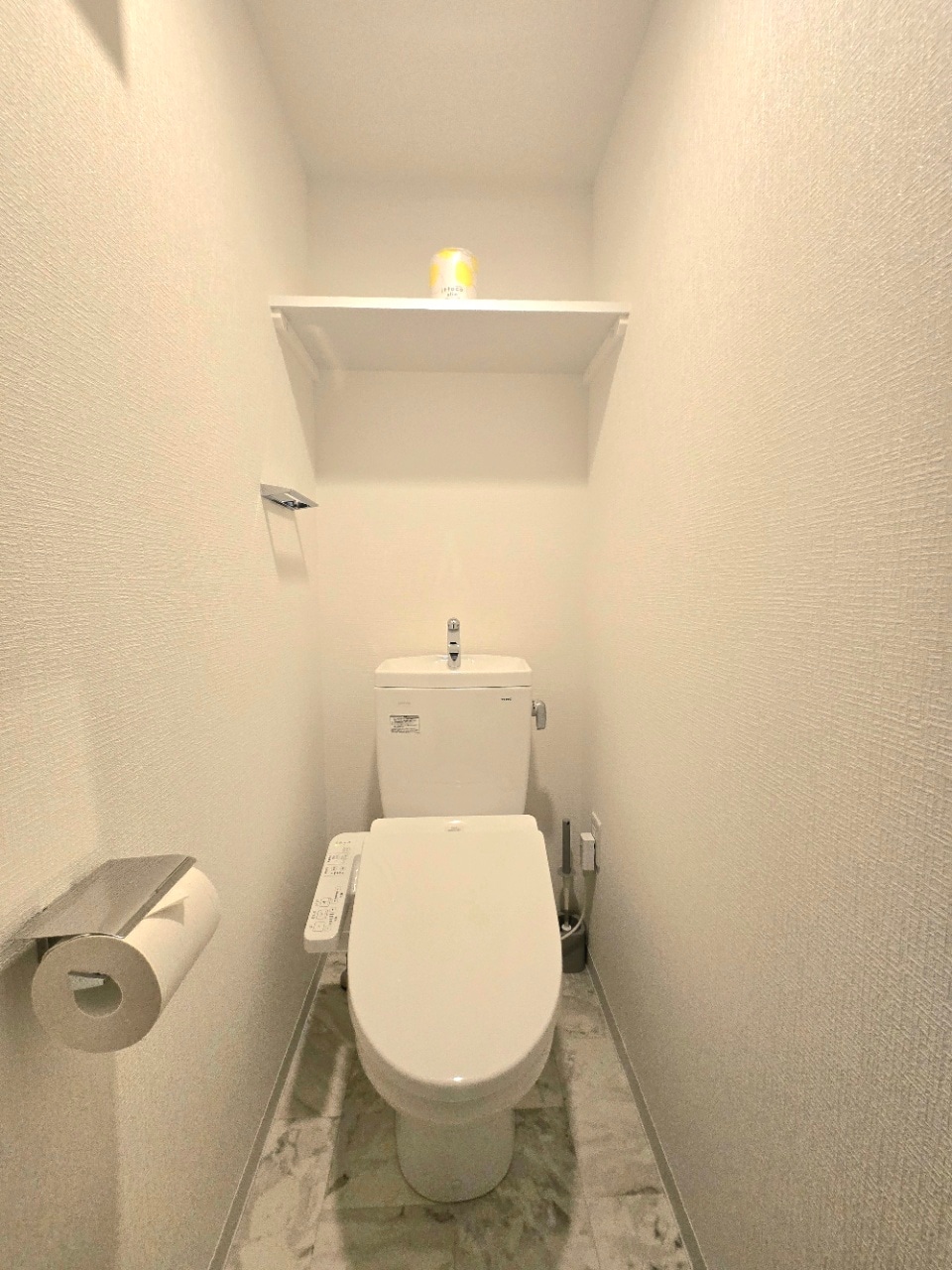 A clean and bright restroom space features a modern toilet positioned against a neutral wall. A small shelf above holds a single item, and a roll of toilet paper is mounted nearby. The floor is tiled with light-colored marble, contributing to the overall simplicity.