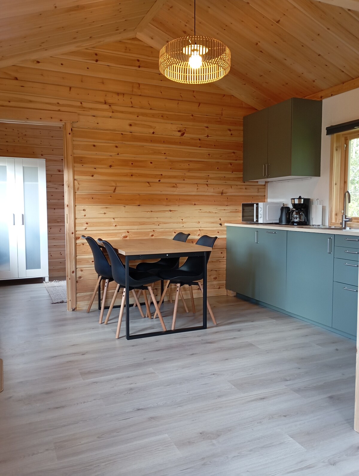 A modern kitchen area features wooden walls and a light wood floor. A black dining table with four chairs is positioned near a large window, allowing for natural light. Green cabinetry is complemented by stainless steel appliances and a coffee maker.