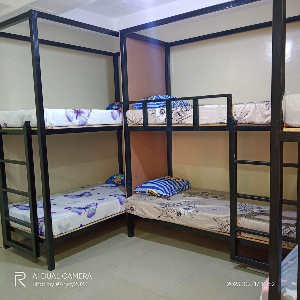 A bunk bed setup is displayed, featuring two double-decker units in a simple design. Each bed is dressed with patterned bedding, including blue and white stripes. The room is brightly lit, with smooth tiled flooring and neutral-colored walls.