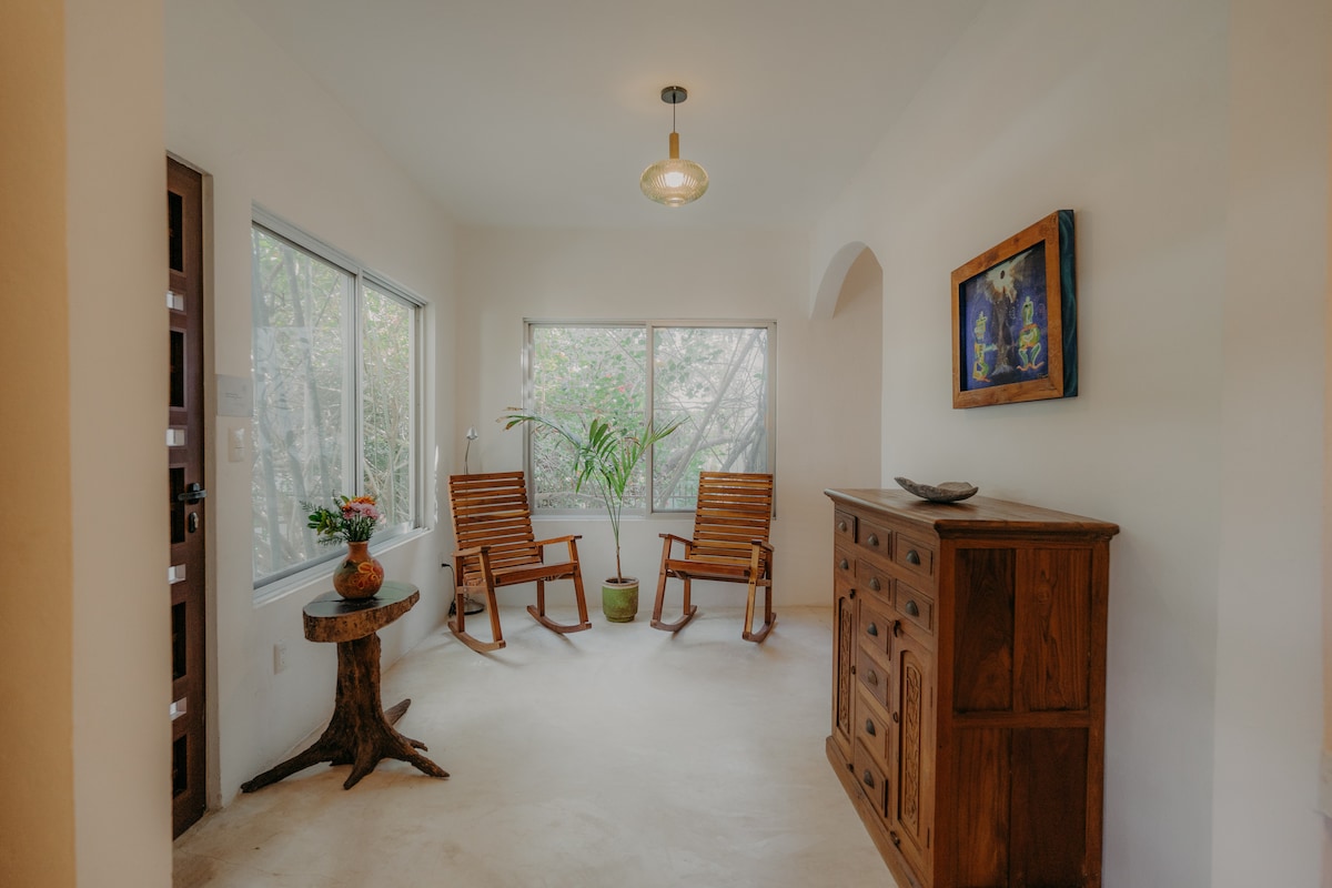 A bright entryway features two wooden rocking chairs positioned by large windows, allowing natural light to filtering in. A potted plant adds a touch of greenery next to the chairs. A rustic wooden cabinet stands against the wall, complementing the serene atmosphere.