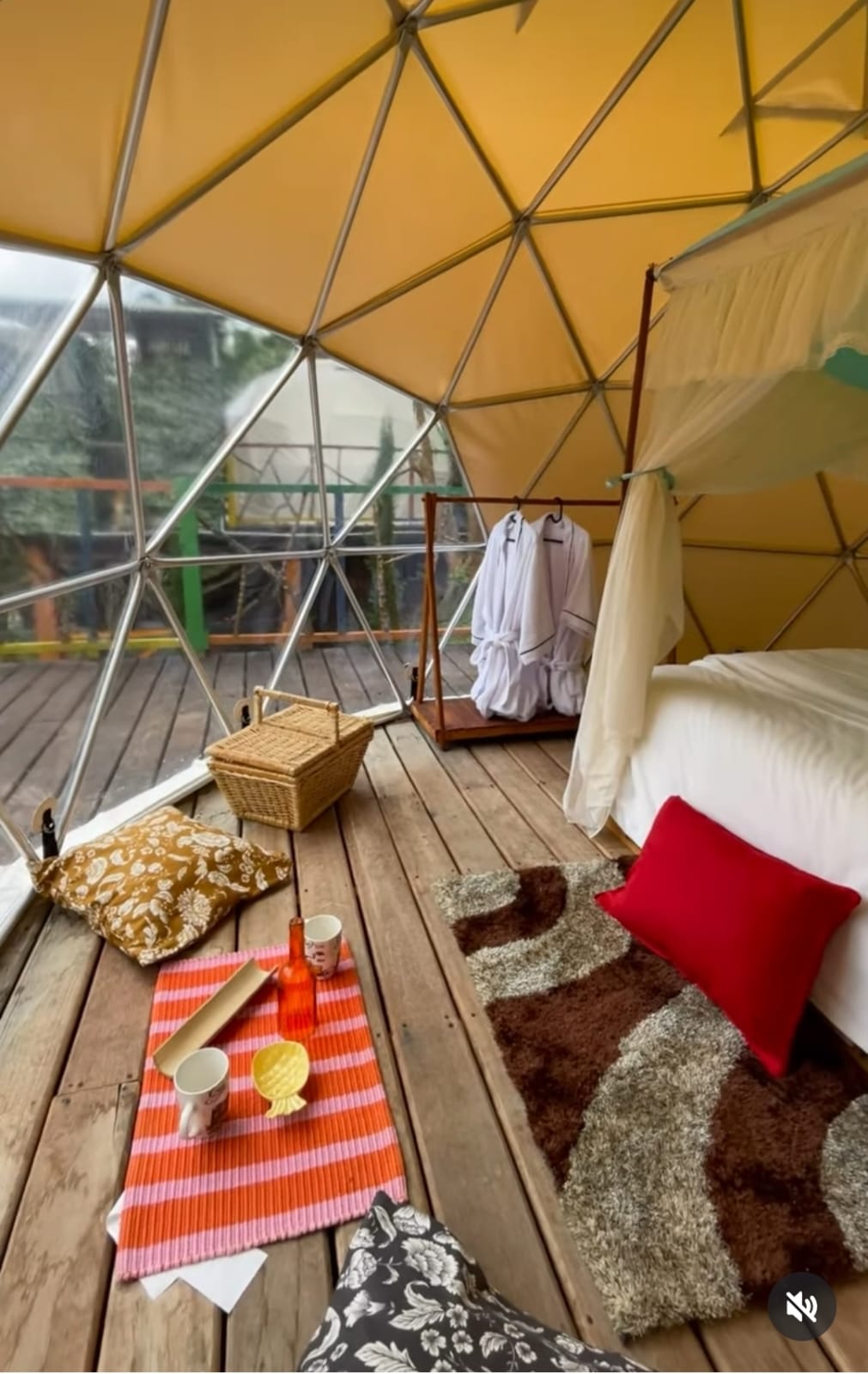An inviting glamping interior features a spacious bed dressed in white linens, surrounded by a warm, patterned rug. A picnic setup with colorful plates and drinks is displayed on a striped mat, while bathrobes hang on a wooden rack, offering a cozy atmosphere.
