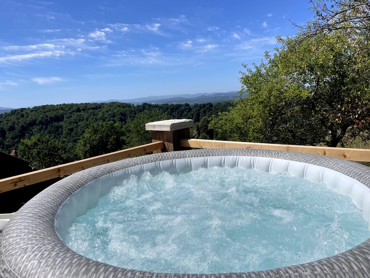 A private jacuzzi is set outdoors, surrounded by wooden decking and nature. Bubbles create a relaxing ambiance in the water, with expansive views of rolling hills and a clear blue sky in the background, providing a serene and inviting atmosphere.