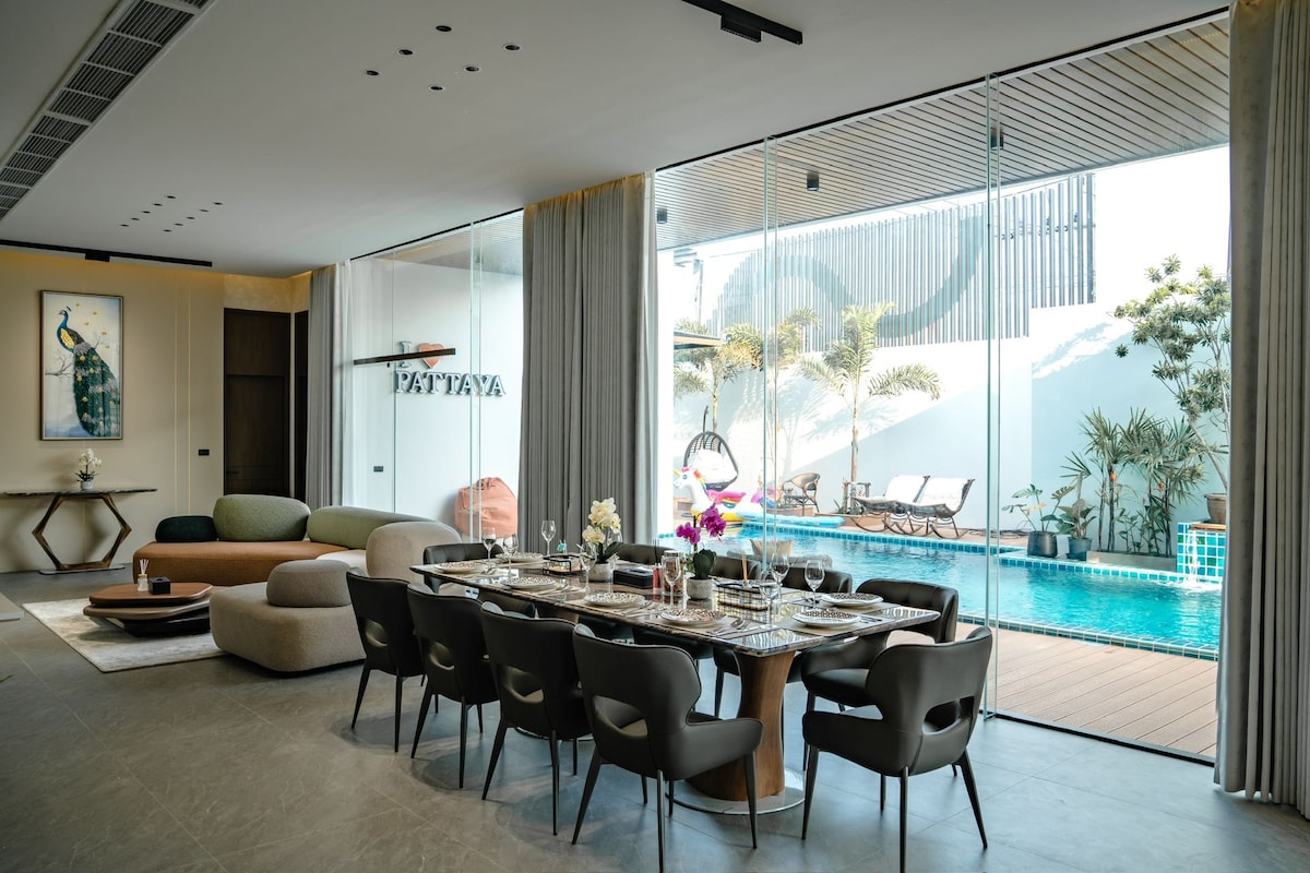 A spacious dining area features a long wooden table set for eight, surrounded by comfortable, modern chairs. Floor-to-ceiling glass walls offer views of the outdoor pool and lounging area, creating a seamless connection between indoor and outdoor spaces.