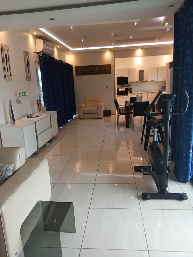 A spacious living area is depicted, featuring glossy tile flooring and modern furnishings. Deep blue curtains frame the large windows, while a comfortable chair and dining table are visible. An exercise bike is positioned near the entrance, leading towards an open kitchen area equipped with appliances.