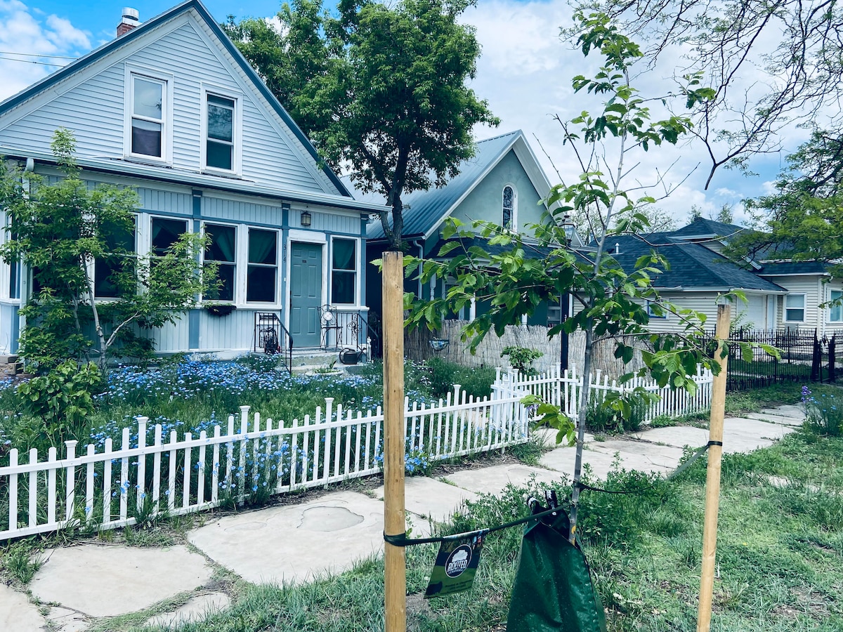 A charming exterior of a two-story house is visible, featuring a front porch and a light blue facade. Lush greenery surrounds the home, with a white picket fence enclosing the landscaped yard filled with blue flowers. The pathway leads to the entrance, inviting guests to explore.