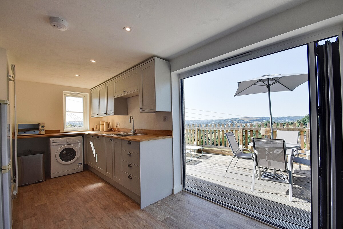 A spacious kitchen features modern grey cabinetry and wood countertops. Bi-fold doors lead to a decking area with outdoor seating under an umbrella, offering views of the surrounding hills. Appliances, including a washing machine, are conveniently integrated into the design.