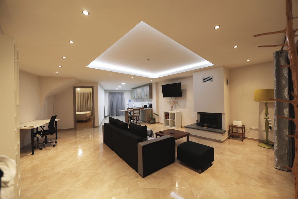 A spacious living area features a black sectional sofa and an ottoman, centered around a coffee table. A wall-mounted television and a contemporary fireplace are set against a light-colored wall. Recessed lighting and natural light illuminate the room, enhancing its inviting feel.